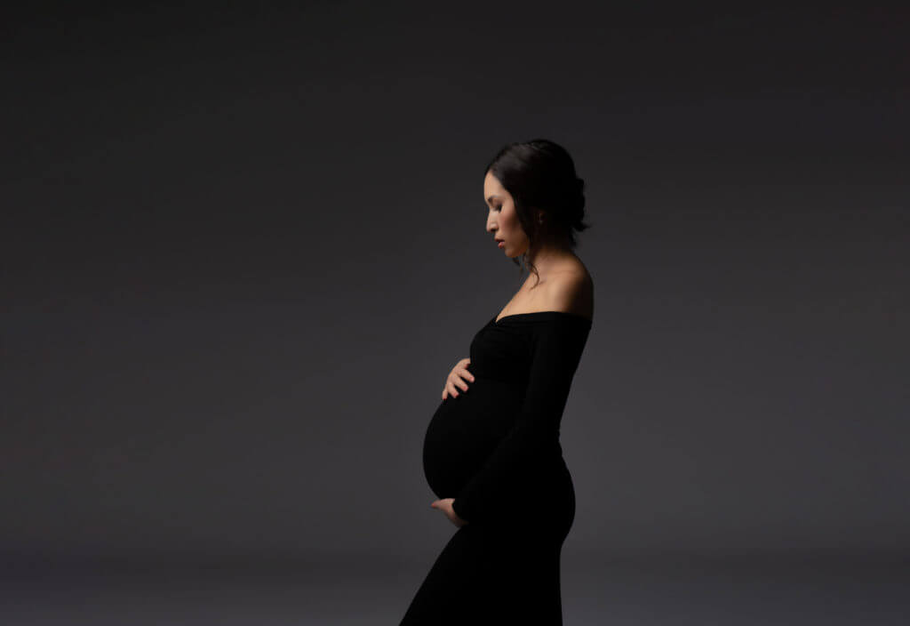 Das Bild zeigt eine schwangere Frau, die selbstbewusst vor einem neutralen grauen Hintergrund posiert. Sie ist elegant in einem schwarzen Kleid gekleidet, das ihre werdende Form betont. Die anmutige Haltung und der gelassene Ausdruck der Frau strahlen ein Gefühl von Ruhe und Erwartung aus.