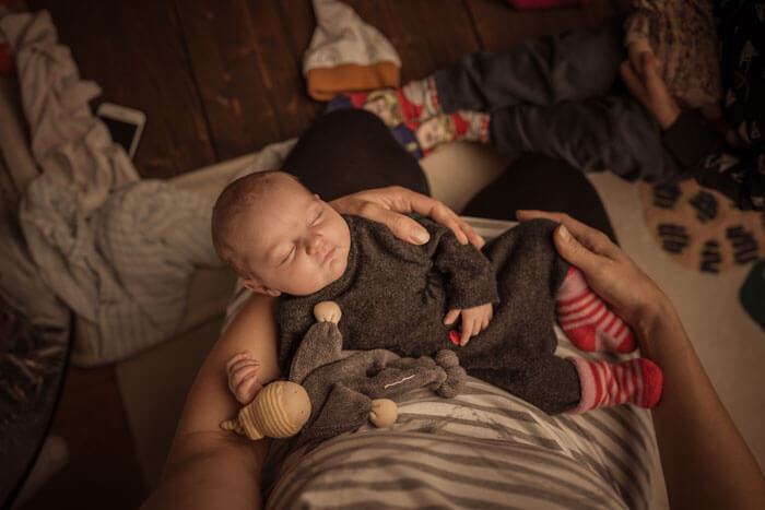 "Eine Frau hält liebevoll ihr Baby in den Armen in einem gemütlich beleuchteten Raum. Im Hintergrund sitzt ein Kind auf einem Holzboden, und in der Nähe ist ein Bett mit einem Teddybären zu sehen, der die familiäre Atmosphäre unterstreicht.