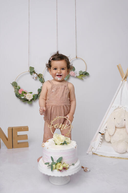 Baby-Mädchen im tan Romper steht vor einem Kuchen mit dem Wort 'one', neben einem weißen Tipi mit einem weißen Hasen-Stofftier und einem Kartonbuchstaben 'N' im Vordergrund.