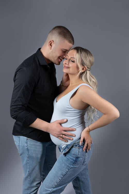 Schwangeres Paar in liebevoller Umarmung vor grauem Hintergrund, erwartungsvoll und freudig auf die Ankunft ihres Kindes wartend.