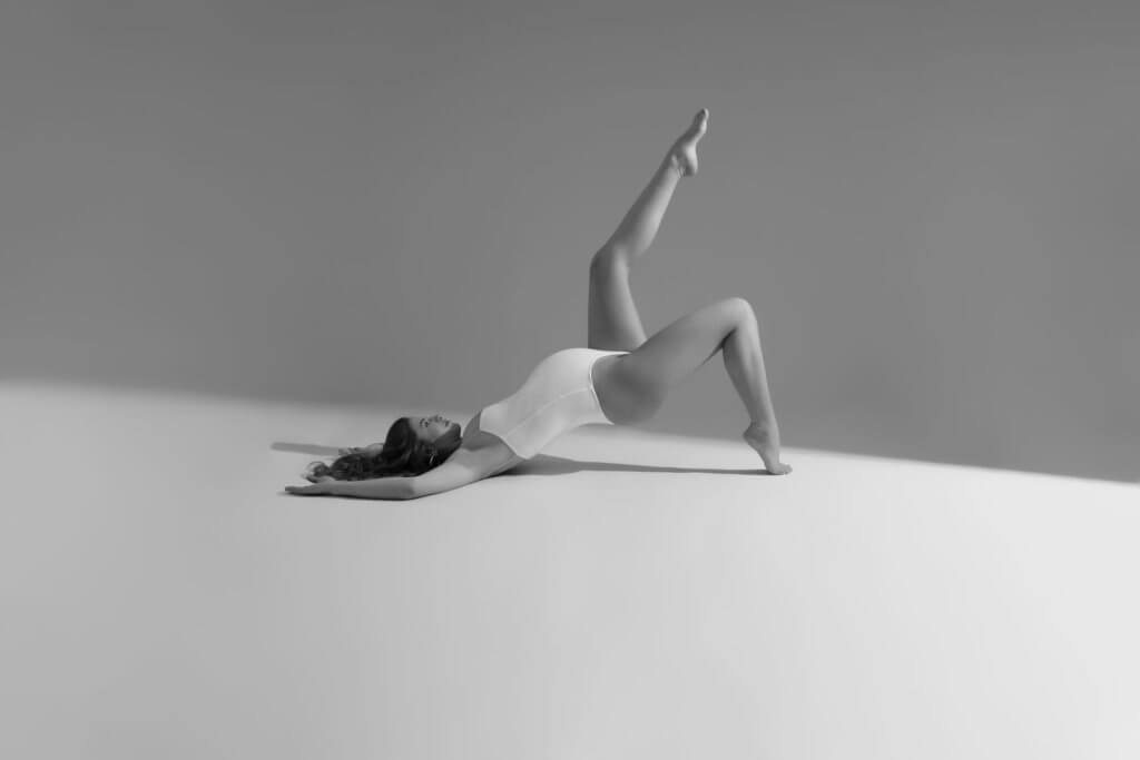 In einer faszinierenden Schwarz-Weiß-Fotografie wird die ätherische Schönheit einer Frau in einer ruhigen und nachdenklichen Pose eingefangen. Liegend auf dem Boden, ruht sie auf ihrem Rücken und strahlt sowohl Verletzlichkeit als auch Stärke aus. Ihre schlanke Figur ist mit einem einfachen, aber eleganten weißen Badeanzug geschmückt, der ihre anmutige Haltung betont.