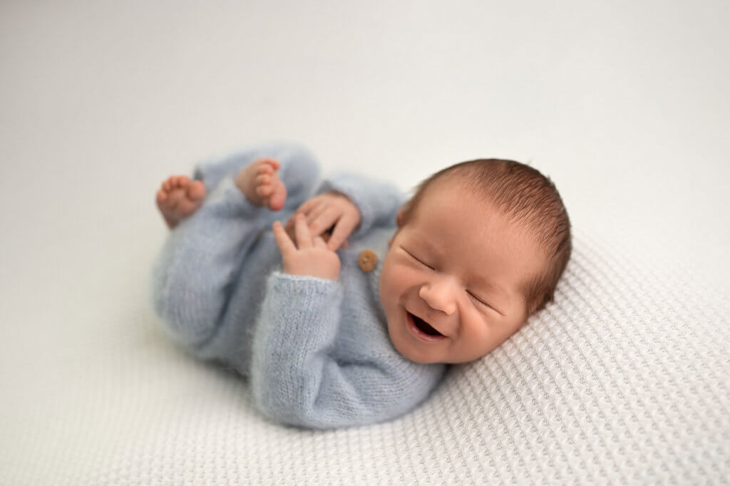 Ein fröhlicher Babyjunge, liegt auf einer weißen Decke und lacht herzlich. Seine rosigen Wangen strahlen vor Freude.